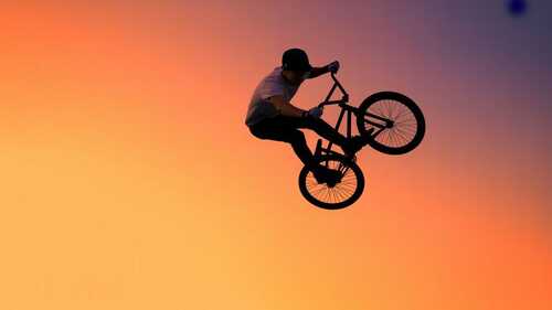 Ciclista de BMX realizando un truco aéreo espectacular en un skatepark con cielo degradado naranja y púrpura