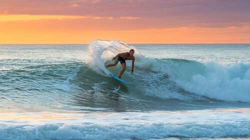 Surfista cabalgando una potente ola en la costa española con atardecer naranja y púrpura de fondo